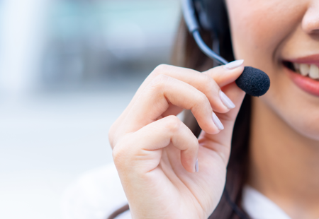 Close up photo showing the lower half of a woman's face. She is smiling and wearing a telephone headset. She is holding the microphone part with her right hand
