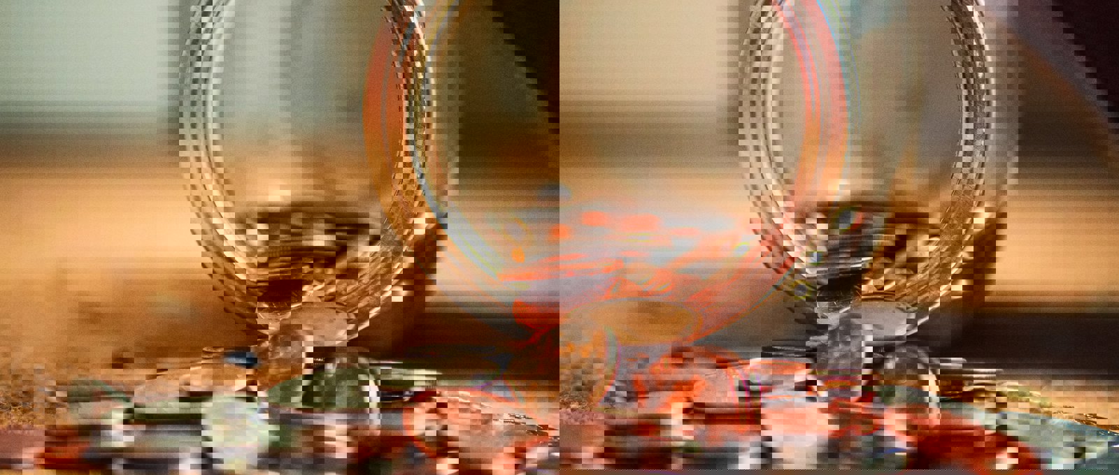 close up photo of a glass jar lying overturned on a surface with lots of coins spilling out of it onto the surface