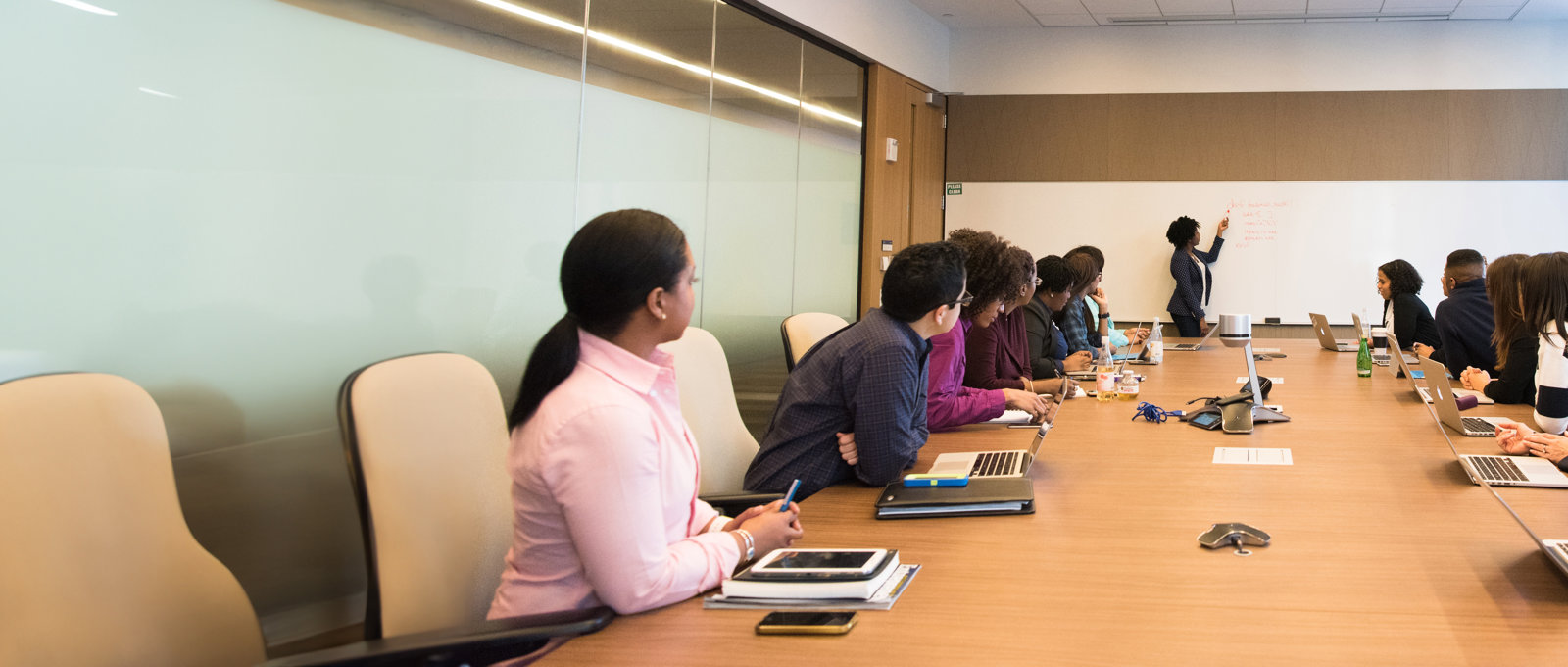 Photograph of several people seated around a long table, each with a laptop in front of them. The photo is taken from a low angle looking along the table. At the far end a woman is writing on a whiteboard