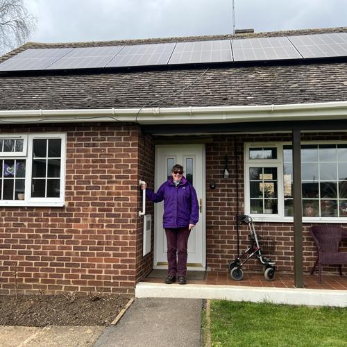 Photo of a Futures' customer called Lucille, standing on the front step outside her bungalow. She's wearing a purple coat and purple trousers, has dark hair tied up and sunglasses. Her bungalow has solar panels on the roof.