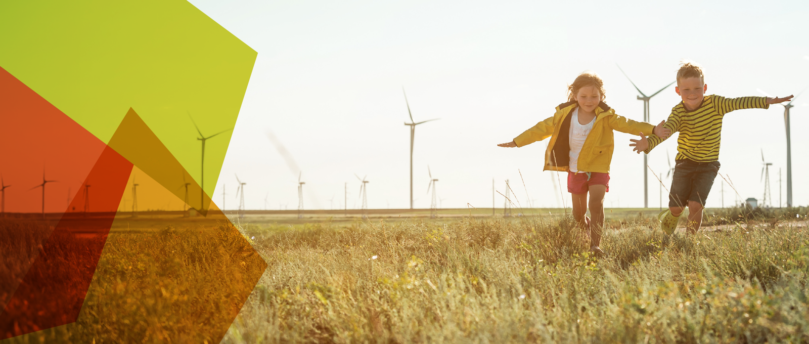 Photo of two happy children holding hands and smiling as they run towards the camera. It is a sunny day and there is a windfarm behind them