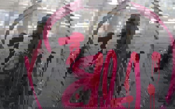Photo of a man in sunglasses and a hoodie spraying red graffiti on glass - viewed from the other side of the glass