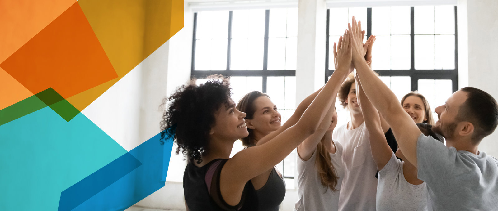 Photo of seven casually dressed young people in what looks like an office environment with a bright window. They are grouped in a circle and have their hands raised up for a group 'high five'